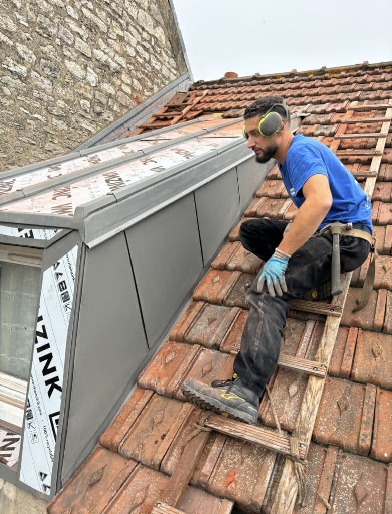 Un couvreur 78 effectuant l'installation d'un bardage en zinc sur une toiture en tuiles, équipé d'une échelle de toit.