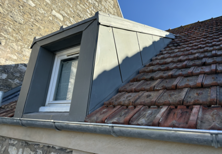 Lucarne rénovée sur une toiture en tuiles anciennes dans les Yvelines, réalisée par un couvreur professionnel. La lucarne est habillée de métal pour une finition moderne, avec un encadrement soigné et des gouttetières en zinc.